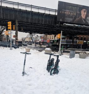 Two Veo micromobility and a scooter are abandoned on a snow-covered sidewalk.